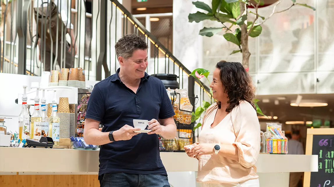 Twee collega's staan vrolijk te praten bij de koffiebar op kantoor.